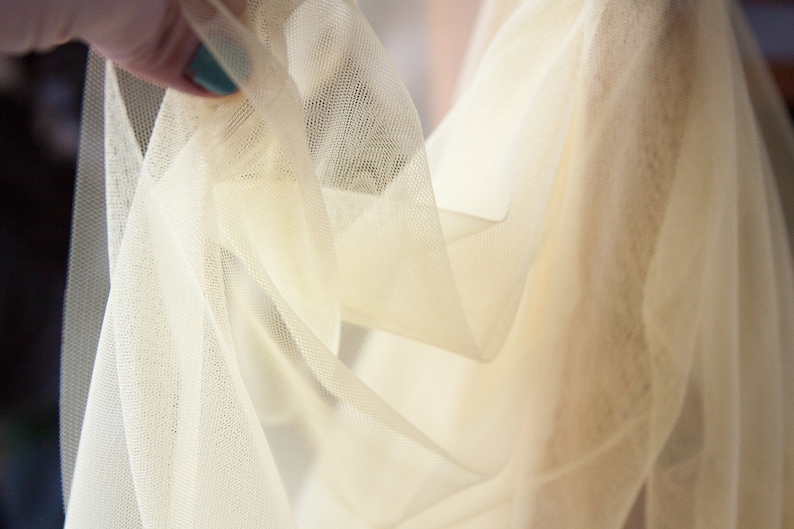 May include: Close-up of a sheer, ivory-colored tulle fabric. The fabric is soft and delicate, with a slightly textured surface.