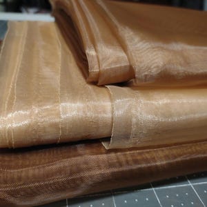 May include: Two pieces of sheer fabric in a light brown color. The fabric is folded and layered on top of each other. The fabric has a subtle shimmer.