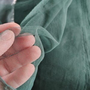 以下が含まれることがあります： 濃いティール色のチュール生地の見本を手に持ったクローズアップ。生地は細かい六角形のメッシュパターンです。背景には、さらに多くの生地とカッティングマットが表示されています。