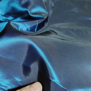 Op de afbeelding: Close-up van iriserende stof in tinten van blauwgroen en marineblauw. De stof heeft een glad, reflecterend oppervlak met zichtbare vouwen en kreukels. Het materiaal lijkt licht en vloeiend, met een subtiele glans. Het algehele effect is er een van diepte en rijkdom.