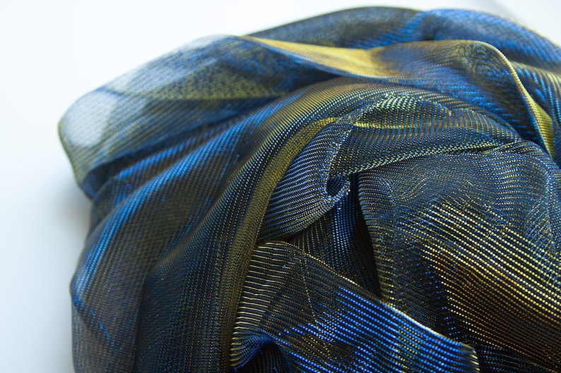 May include: A close-up of a blue and gold metallic fabric with a shiny, iridescent finish. The fabric is folded and draped, creating a textured and abstract pattern.