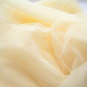 May include: A close-up of a pale yellow tulle fabric, showing its delicate texture and soft, flowing drape.