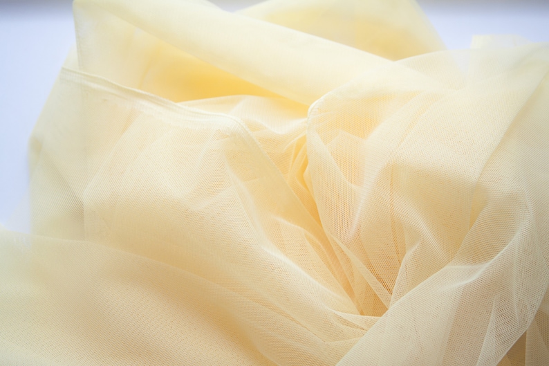 May include: A close-up of a pale yellow tulle fabric, showing its delicate texture and soft, flowing drape.