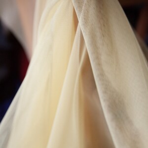 May include: A close-up of a sheer, ivory-colored fabric with a delicate, woven texture. The fabric is draped and folded, creating soft, flowing lines.