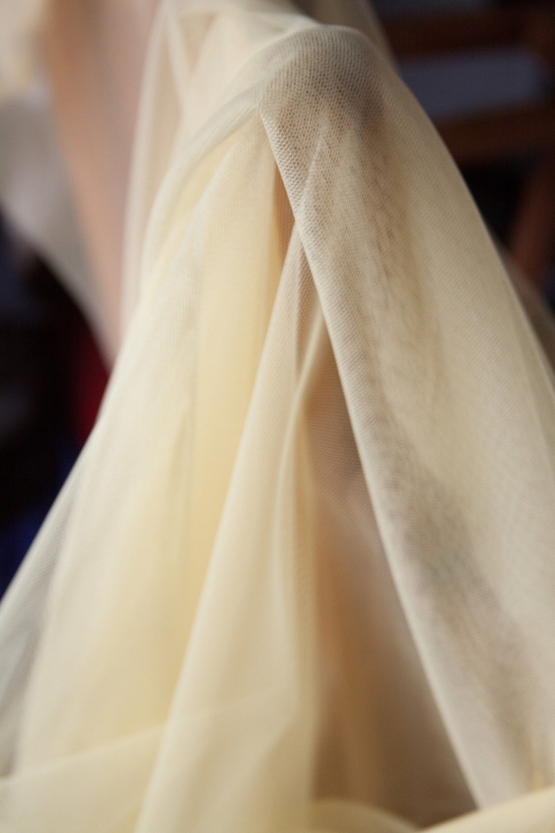 May include: A close-up of a sheer, ivory-colored fabric with a delicate, woven texture. The fabric is draped and folded, creating soft, flowing lines.