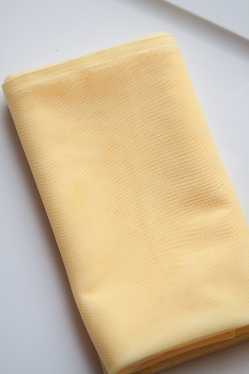 May include: A pale yellow tulle fabric, folded and displayed on a white surface.
