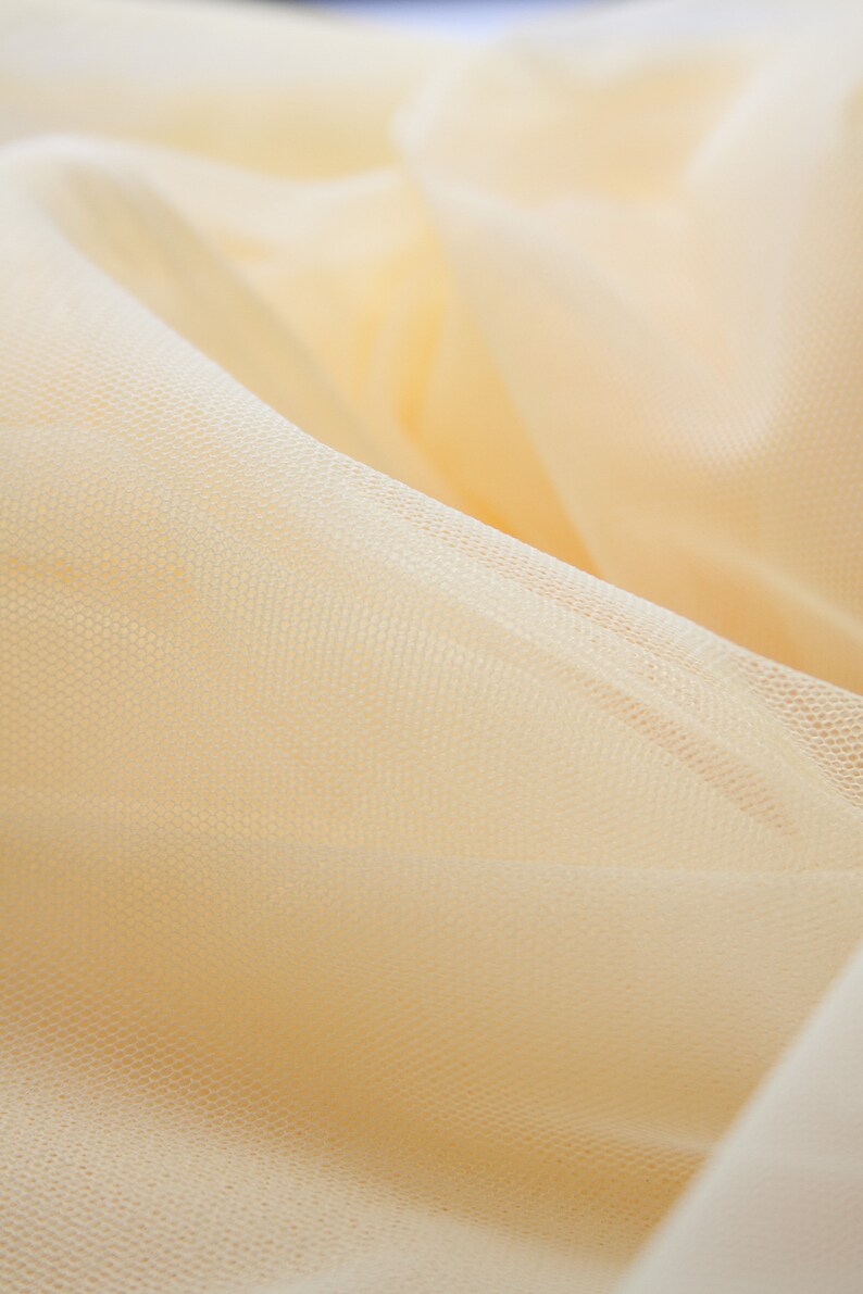 May include: Close-up of a pale yellow tulle fabric with a fine mesh texture. The fabric is soft and delicate, with a subtle sheen.