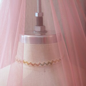 May include: A pale pink tulle fabric draped over a dress form. The fabric is sheer and has a delicate texture. The dress form is made of a light-coloured material and has a simple design.