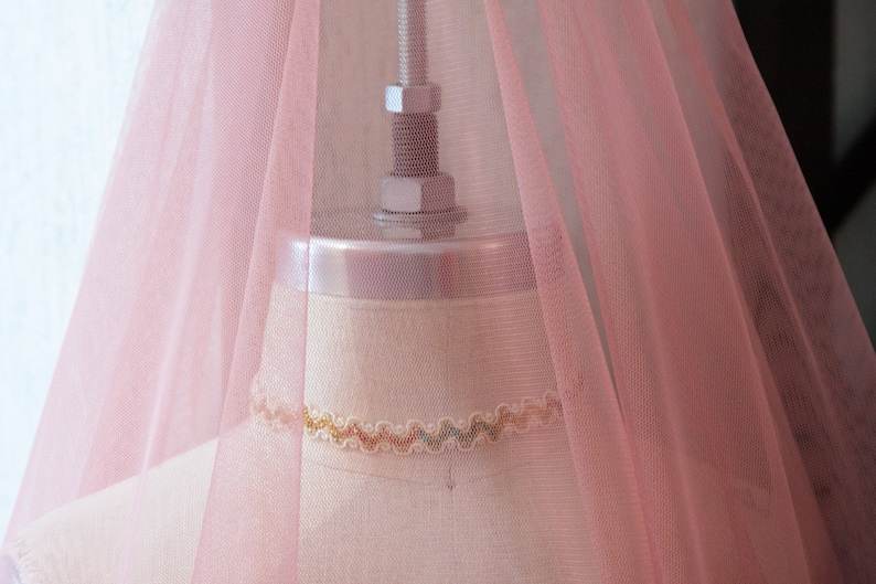 May include: A pale pink tulle fabric draped over a dress form. The fabric is sheer and has a delicate texture. The dress form is made of a light-coloured material and has a simple design.