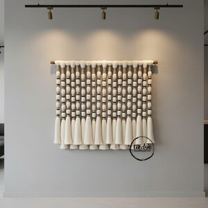 May include: A cream-colored, handmade wall hanging with a wooden dowel. The design features rows of white beads and tassels, with dark brown accents. The bottom of the piece has a series of long, white tassels. The text "tiedye handmade art" is in a circle on the bottom right.