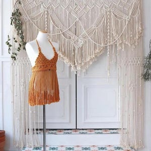 May include: A handmade macrame wall hanging and a halter top dress. The wall hanging is cream-colored with intricate knotting and fringe. The halter top dress is a golden-brown color with a halter top and fringe skirt. The items are displayed in front of a white door.