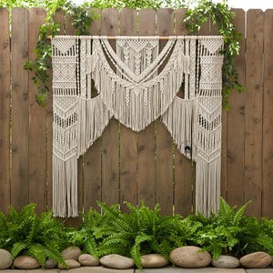 May include: A cream-colored macrame wall hanging with intricate geometric patterns and fringe. The textile is suspended from a wooden dowel against a wooden fence, with greenery and ferns in the foreground. This decorative piece adds a bohemian touch to any space.