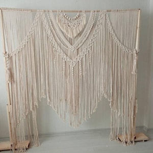 May include: A large, cream-colored macrame wall hanging with a wooden frame. The design features intricate knotting patterns, including diamond shapes and cascading fringe. The piece is displayed on a wooden stand, creating a bohemian aesthetic.