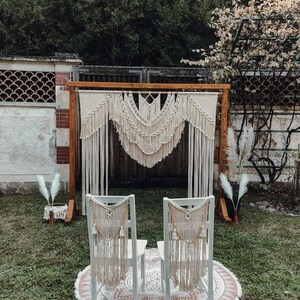 May include: A bohemian-style wedding ceremony setup featuring a wooden arch adorned with cream-colored macrame. Two white chairs with macrame backdrops are set on a round rug. The scene is outdoors with greenery in the background.
