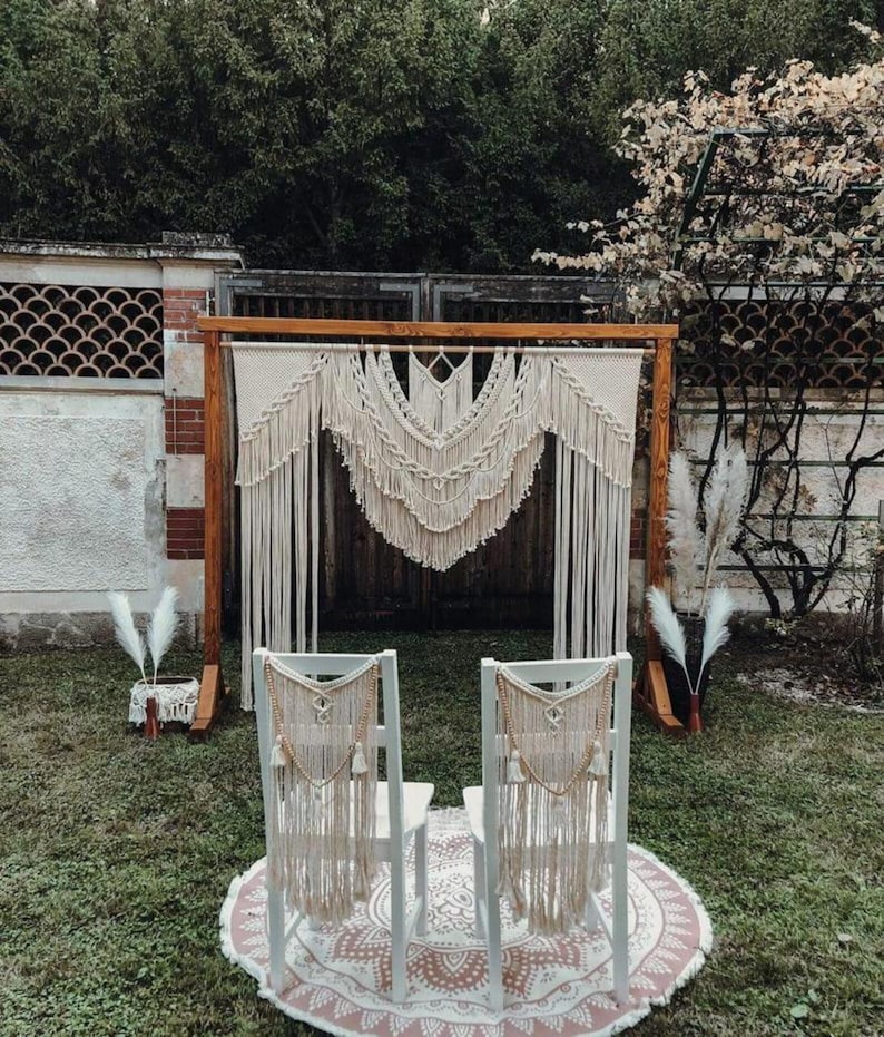 May include: A bohemian-style wedding ceremony setup featuring a wooden arch adorned with cream-colored macrame. Two white chairs with macrame backdrops are set on a round rug. The scene is outdoors with greenery in the background.