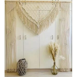 May include: A cream-coloured macrame wall hanging with a geometric design, featuring diamond and fringe details. The macrame is suspended from a wooden rod. A woven basket and a glass vase with dried flowers are in the foreground.