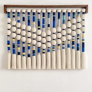 May include: A decorative wall hanging featuring a series of vertical strands. Each strand has a white tassel at the bottom, a series of white spheres, and sections of blue, yellow, and gray stripes. The piece is mounted on a wooden frame.