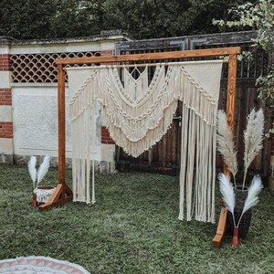 May include: A wooden wedding arch with a macrame backdrop. The arch is made of light brown wood and features a cream-colored macrame design with fringe. Decorative pampas grass is placed on either side of the arch, creating a bohemian aesthetic.
