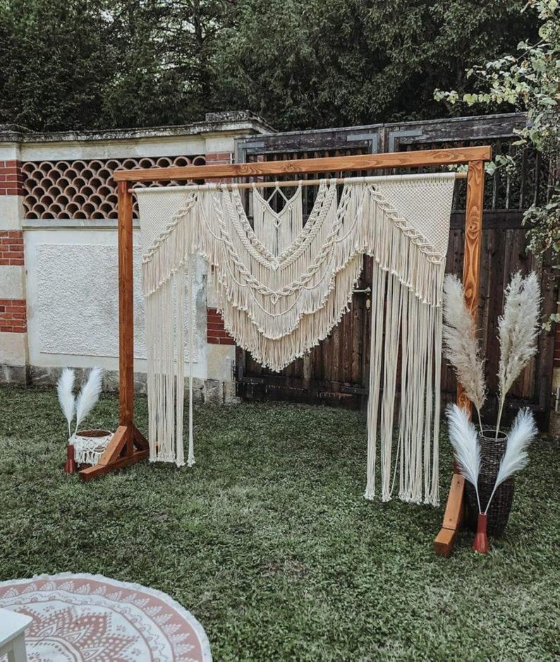 May include: A wooden wedding arch with a macrame backdrop. The arch is made of light brown wood and features a cream-colored macrame design with fringe. Decorative pampas grass is placed on either side of the arch, creating a bohemian aesthetic.