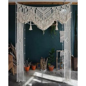 May include: Cream macrame wall hanging with an arched valance and vertical panels. The macrame features intricate knotting patterns and fringe details. The bohemian-style decor is displayed against a dark teal wall.