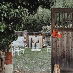 May include: An outdoor wedding ceremony setup with white chairs, a macrame backdrop, and decorative orange paper fans. The scene is set in a grassy yard with a wooden gate and surrounding trees.
