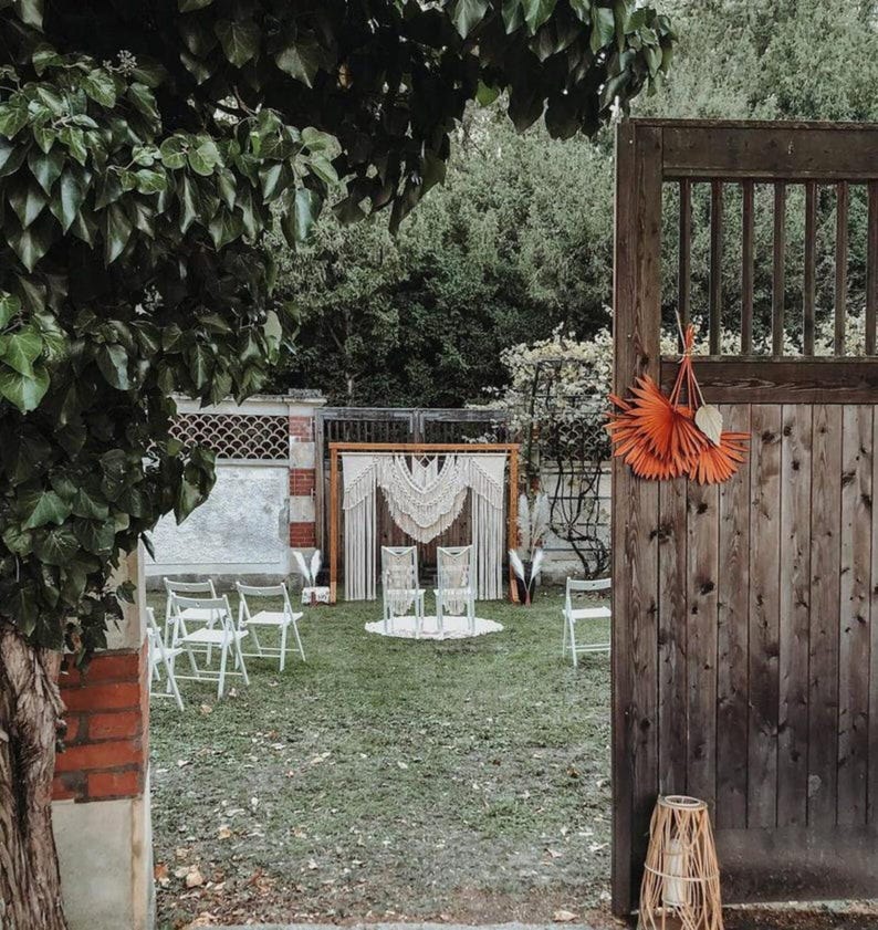 May include: An outdoor wedding ceremony setup with white chairs, a macrame backdrop, and decorative orange paper fans. The scene is set in a grassy yard with a wooden gate and surrounding trees.