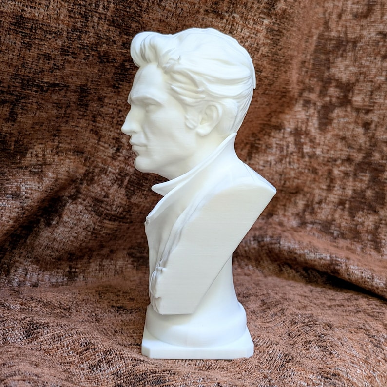 May include: A white bust sculpture depicting a person in profile with styled hair and a high-collared garment. The figure features defined facial contours, a sharp jawline, and a smooth matte finish, presented as a solid, monochromatic head and shoulder piece.