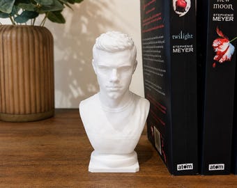 May include: A white matte bust sculpture of a young person with short hair, positioned between two black books titled Twilight and New Moon by Stephenie Meyer. The sculpture features a smooth finish and a round base, capturing a neutral facial expression.
