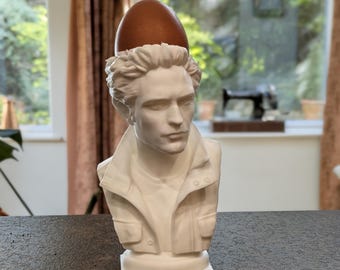 May include: A white 3D printed bust of a person with short hair wearing a collared jacket, featuring a brown egg resting on top of the head. The sculpture is designed as an egg cup, providing a stable base for the egg to sit securely.