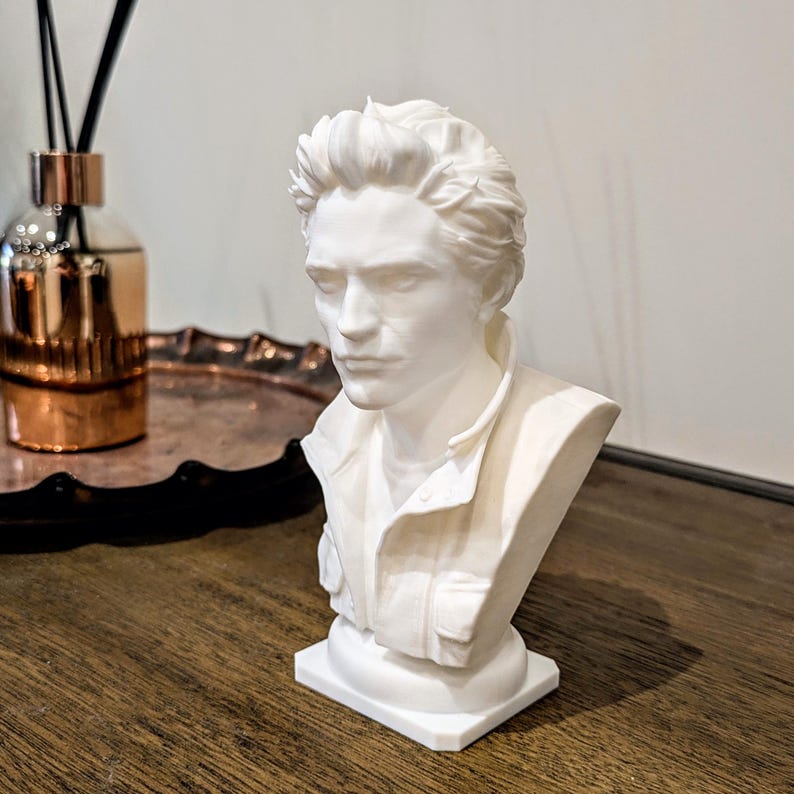 May include: A white matte bust sculpture depicting a person with short, textured hair and a collared jacket. The figure has a neutral facial expression, defined features, and is mounted on a rectangular base, presented as a solid monochromatic object.