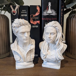 May include: Two off-white, speckled stone-effect busts depicting a man with short, textured hair and a woman with long, flowing hair. The figures are positioned in front of a row of Twilight book series novels, including Twilight, New Moon, Eclipse, and Breaking Dawn.