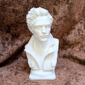 May include: A white bust sculpture depicting a person with short, textured hair, wearing a collared jacket with visible buttons and pockets. The figure has a neutral facial expression with eyes closed, presented as a solid, monochromatic piece.