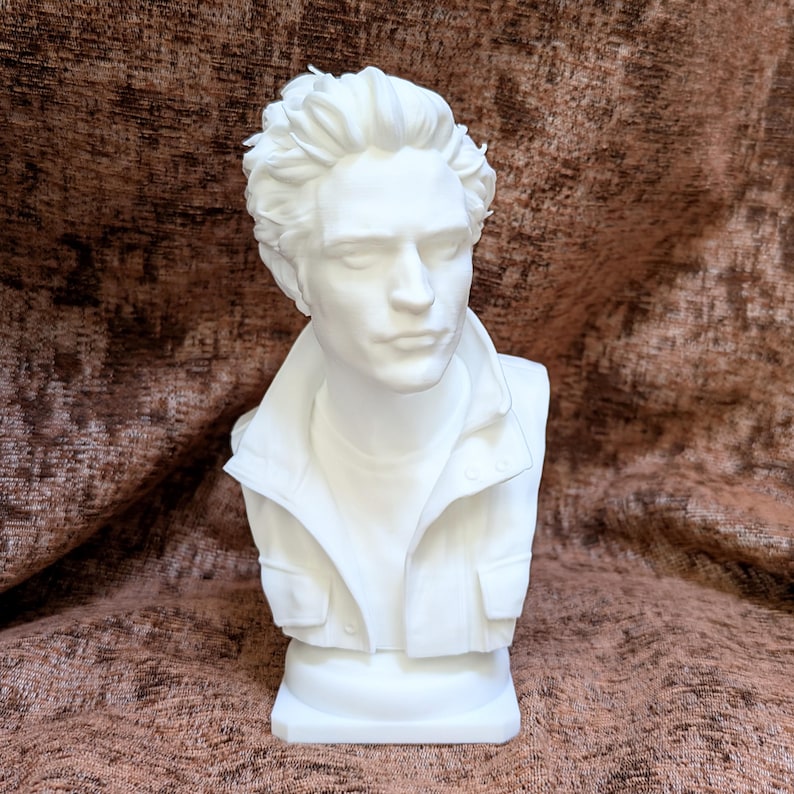 May include: A white bust sculpture depicting a person with short, textured hair, wearing a collared jacket with visible buttons and pockets. The figure has a neutral facial expression with eyes closed, presented as a solid, monochromatic piece.