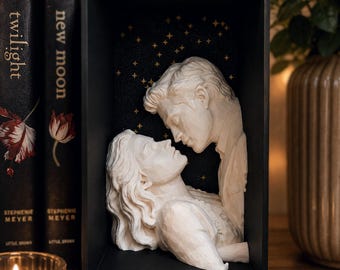 May include: A white sculpted bust of two figures facing each other in a romantic pose, housed inside a black rectangular shadow box. The interior back panel of the box is black with small gold star accents, creating a night sky effect.