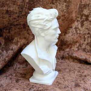May include: A white bust sculpture depicting a person with short, textured hair, shown in profile. The figure wears a collared jacket with visible pockets over a simple shirt, captured from the chest up on a solid, smooth base.