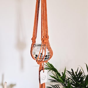 May include: A hanging macrame plant holder in a warm orange hue. It features a glass disco ball planter. The macrame design includes a knotted structure and a tassel. The plant holder is suspended against a white wall with green plants.