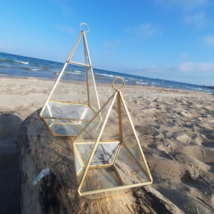 以下が含まれることがあります： 風化した丸太の上に置かれた、金色の幾何学的なガラス製のテラリウムが2つ。テラリウムはピラミッド型で、金色の金属フレームと透明なガラスパネルで構成されています。大きなテラリウムは背景に、小さなテラリウムは前景にあります。