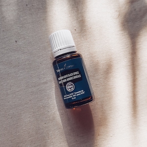 Puede incluir: Una pequeña botella de vidrio oscuro de aceite esencial Young Living Northern Lights Black Spruce con tapa blanca. La etiqueta es de color verde azulado oscuro con texto blanco, incluyendo "100% Pure, YLTG Essential Oil" y "15 ml". La botella está sobre tela beige.