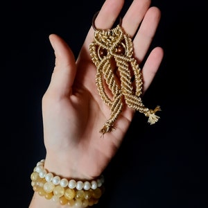 May include: A gold macrame owl keychain with brown wooden beads. The keychain is held in a hand wearing a multi-layered bracelet with white and yellow beads.