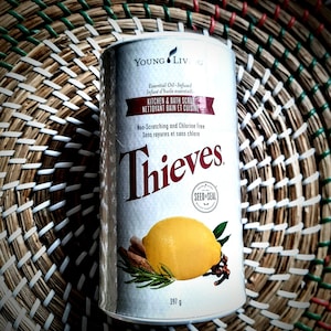 May include: A cylindrical container of "Thieves" kitchen and bath scrub. The label includes text in English and French, a lemon, cinnamon sticks, and rosemary sprigs. The container is 397 g.