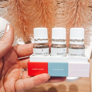 Może przedstawiać: Trzy małe szklane butelki z białymi zakrętkami i brązową cieczą w środku. Butelki są oznaczone napisem "SIMPLIFIED" i ułożone w pudełku z etykietami "CANDY CANE", "WINTER SPA" i "FIREPLACE" z przodu.