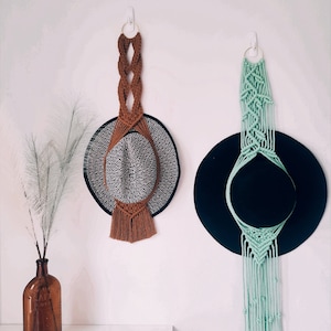 以下が含まれることがあります： 白い壁に白いフックで吊るされた、フリンジ付きの2つのマクラメ製の帽子掛け。1つは茶色、もう1つは緑。茶色のマクラメ製の帽子掛けには白黒の帽子が、緑のマクラメ製の帽子掛けには黒い帽子が掛けられています。