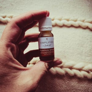 Può includere: Un piccolo flacone di vetro marrone con un tappo bianco e un'etichetta bianca. L'etichetta riporta "SIMPLIFIED by Just Kit" e "PUMPKIN SPICE Premium Essential Oil Blend" con una misura di 0,34 fl. oz. (10 mL).