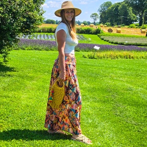 May include: A woman wearing a straw hat, white top, and a long, colorful skirt with a floral pattern. She is holding a yellow crocheted bag. The background features a green field, trees, and a blue sky.
