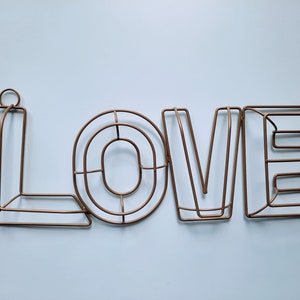 Op de afbeelding: Een metalen draadbeeld van het woord "LOVE" in koperkleur. De letters zijn met elkaar verbonden en hebben een eenvoudig, modern ontwerp.