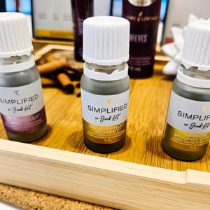 May include: Three small glass bottles with white caps and labels, arranged on a wooden tray. The labels read "Simplified by Jacob Katz" with different scent names: Winter Lily, Grapefruit Sorbet, and Lemon Pound Cake. The bottles contain a dark liquid.