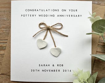 May include: A white card with the text "Congratulations on your Pottery Wedding Anniversary" and "Sarah & Rob 20th November 2016". The card has a brown twine bow with two white clay hearts hanging from it.