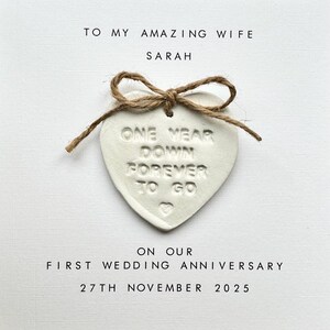 May include: White heart-shaped ornament with the text "One Year Down Forever To Go" tied with twine. The ornament is on a white card with the text "To My Amazing Wife Sarah On Our First Wedding Anniversary 27th November 2025".