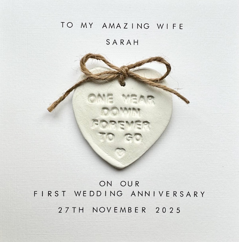 May include: White heart-shaped ornament with the text "One Year Down Forever To Go" tied with twine. The ornament is on a white card with the text "To My Amazing Wife Sarah On Our First Wedding Anniversary 27th November 2025".