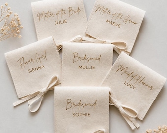 Könnte beinhalten: Cremefarbene Stoffbeutel mit goldfarbenen Aufschriften und Schleifen. Auf jedem Beutel stehen die Namen und Rollen der Hochzeitsgesellschaft: "Mother of the Bride JULIE", "Mother of the Groom MAEVE", "Flower Girl SIENNA", "Bridesmaid MOLLIE", "Maid of Honour LUCY", und "Bridesmaid SOPHIE".